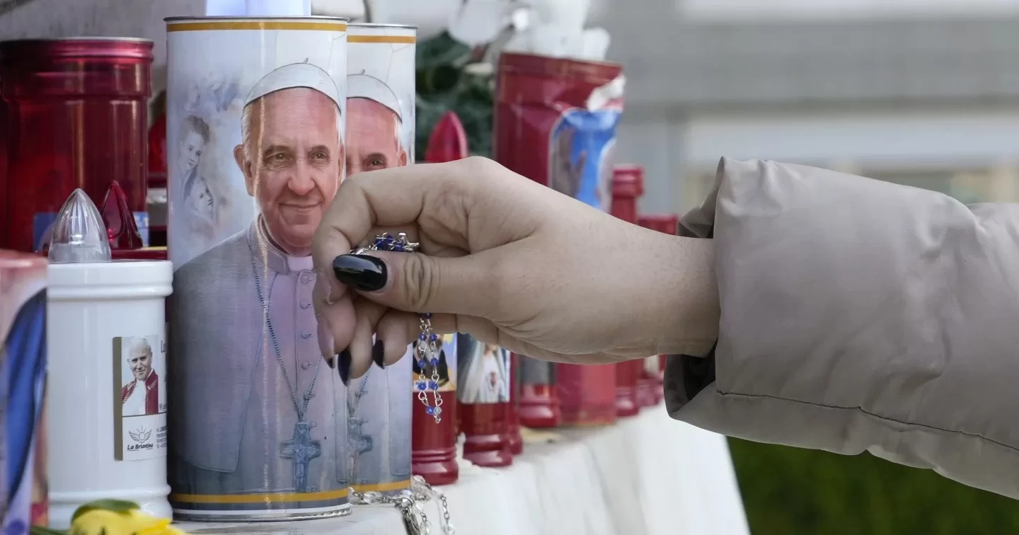 Жінка кладе вервицю біля свічок, прикрашених фотографіями Папи Франциска, біля поліклініки «Аґостіно Ґемеллі», де Папа Франциск бореться з пневмонією, в Римі, субота, 22 лютого 2025 / Фото AP Photo